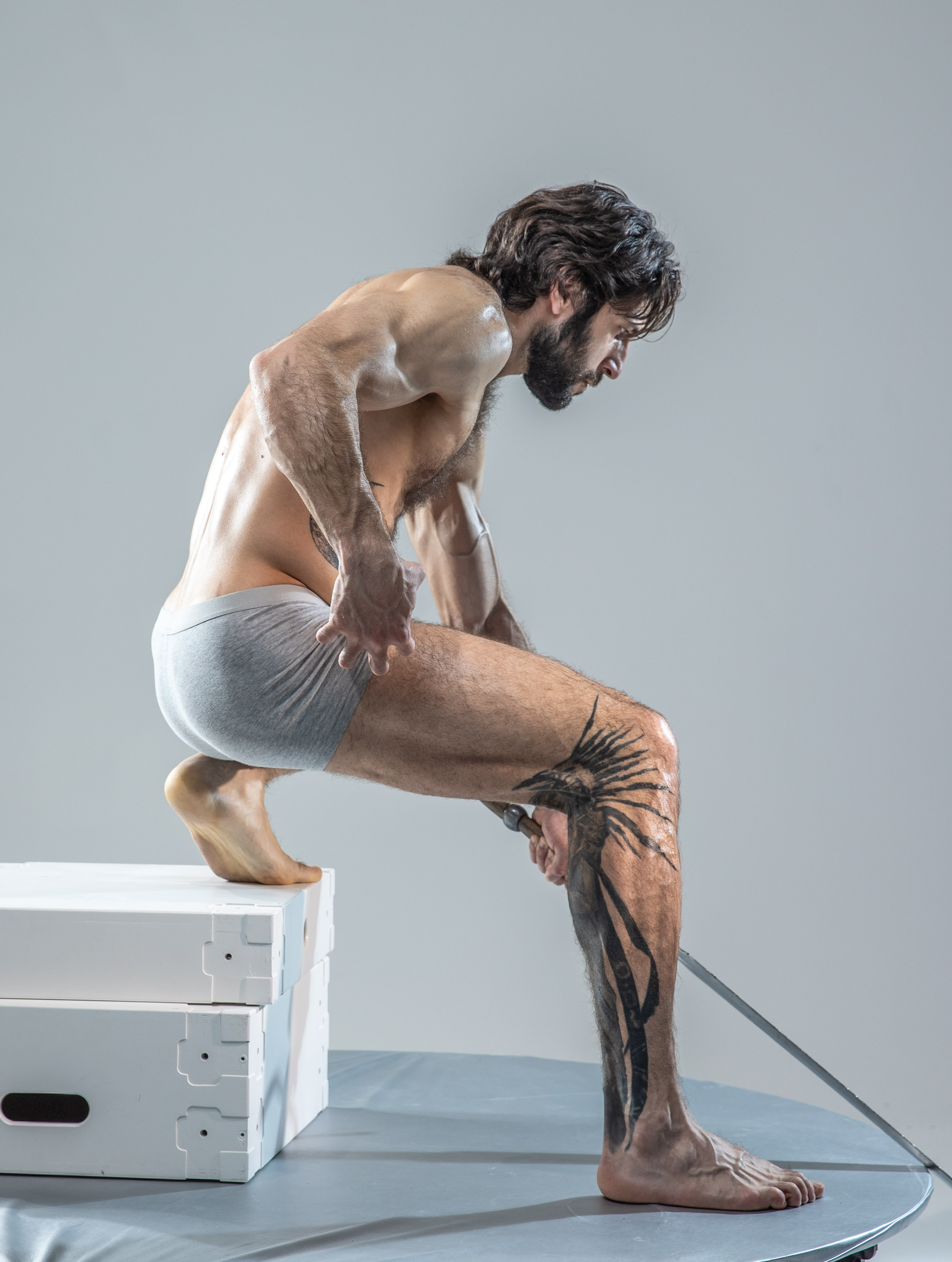 a man in grey underwear is captured midpose in a studio. he's perched on white blocks, one foot raised, and his body angled as if drawing a sword from a sheath on his left leg. the background and floor are a muted gray, focusing attention on the man's form and the dynamic pose.