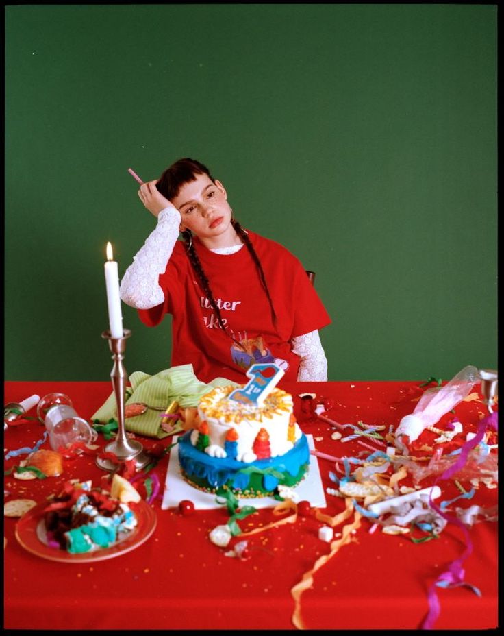 a young girl with red hair is sitting at a table with a messy birthday cake in front of her. she is wearing a red shirt that says "butter like?" the table is covered in a red tablecloth and there is a green wall in the background. the cake is decorated with colorful frosting and sprinkles. it is surrounded by ribbons, streamers, and other party decorations. there are also glasses, an apple, and a plate with a small cake on it. the girl looks bored and sad.