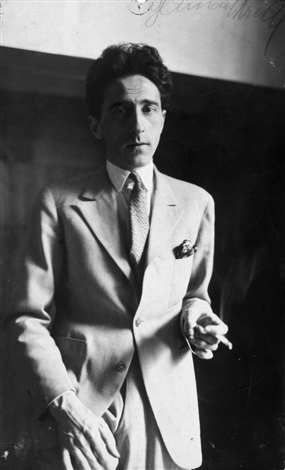 a black and white portrait photograph of a man in a lightcolored suit. he is wearing a tie and appears to be holding a cigarette. the photo is likely from the 1920s or 1930s, judging by the style of clothing and the photographic technique. the man's expression is serious, and he is posed in a formal manner, suggesting a studio portrait. the background is dark and unfocused, drawing attention to the subject.  there is what looks like an inscription or signature in the upperright corner of the image.