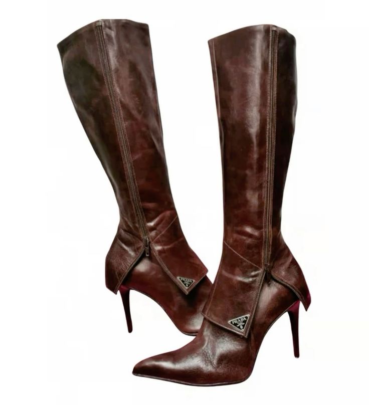 a pair of prada brown leather kneehigh boots with stiletto heels are shown. the boots feature a unique folded leather detail on the sides and a zip closure.  they are positioned on a white background with one boot angled slightly behind the other, showcasing both the front and the side profiles. the prada logo is visible on the folded leather accents.