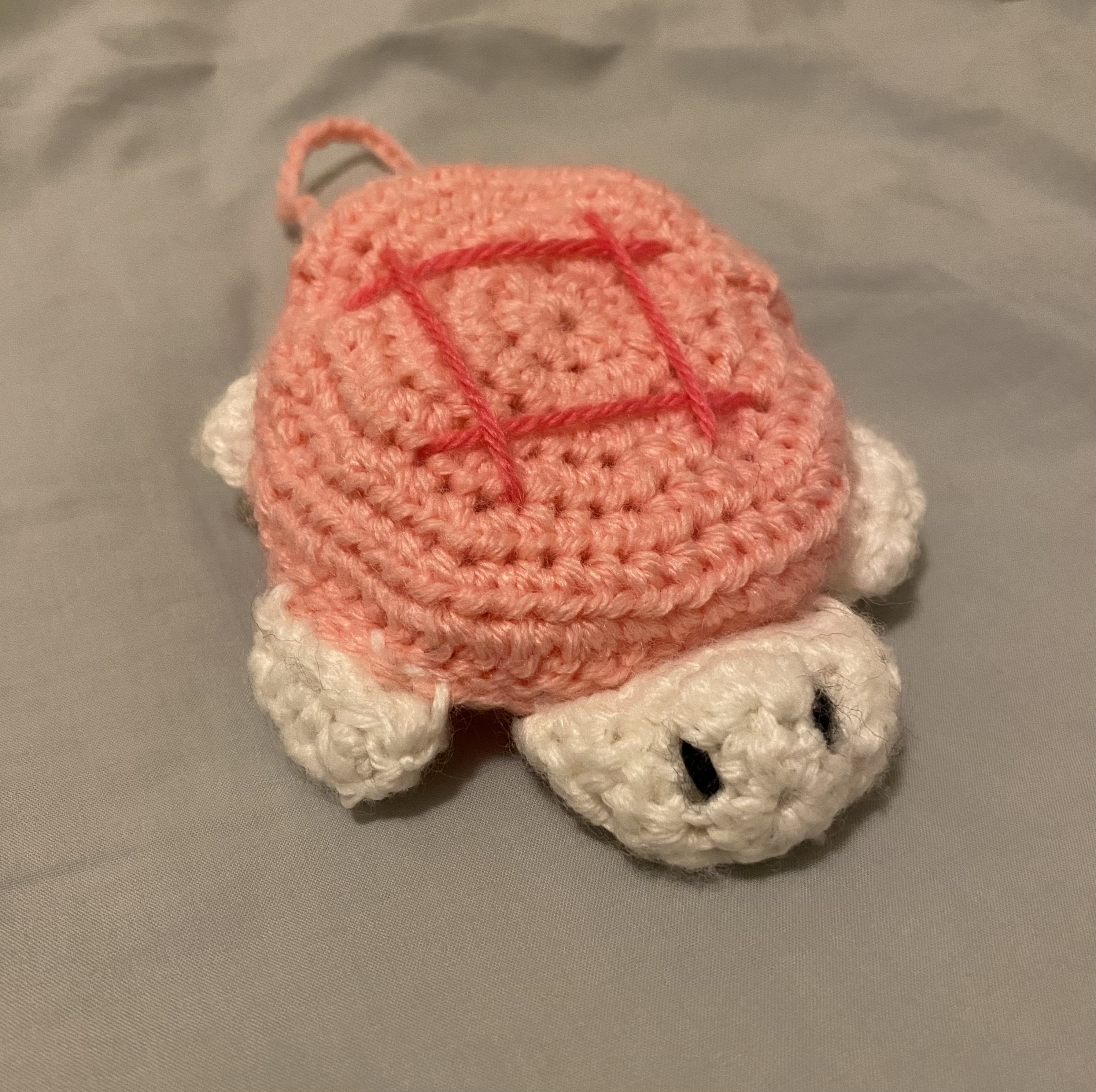 a closeup shot of a crocheted turtle toy. the turtle is predominantly pink with white accents on its feet and head.  a pink cross is stitched onto its shell. the turtle appears small and is likely handmade. the background is a muted gray fabric. the overall style is cute and kawaii.