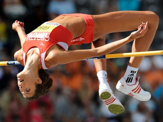 a female athlete clears a high jump bar, in midair, in a competition.  she is wearing a white and red athletic uniform and white sneakers.  her hair is brown, and she is wearing a white socks with a red stripe, with adidas logo. her face is visible.  there are other athletes and spectators in the background, as well as a stadium.