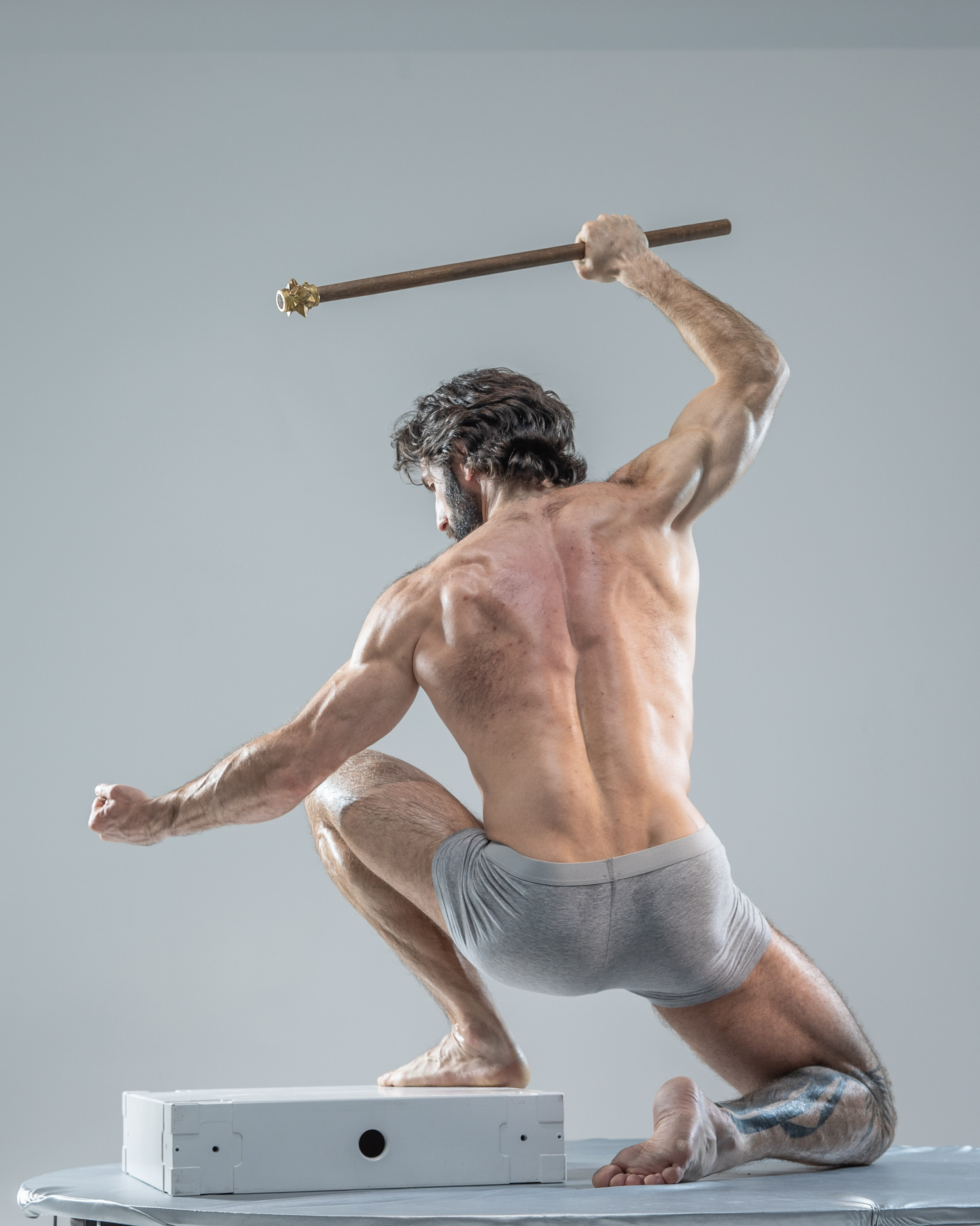 a muscular man with long hair and a beard is captured in a dynamic pose against a grey studio background. he's holding a stafflike object, possibly made of wood with metal accents, suggesting a warrior or mythological figure.  he is wearing only grey underwear, highlighting his physique. the image focuses on the power and athleticism of the male form. the low camera angle enhances the feeling of strength and dynamism.