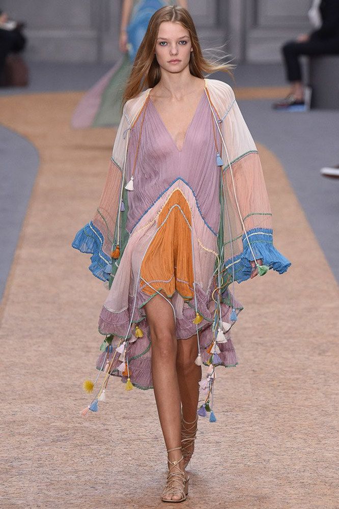 a female model is walking the runway in a colorful, bohemianinspired dress, with long, flowing hair and sandals. she looks chic and glamorous as she struts down the catwalk. the dress is a mix of purple, orange, blue, and green, and is adorned with tassels. the model's long hair falls over her shoulders, and her makeup is minimal.