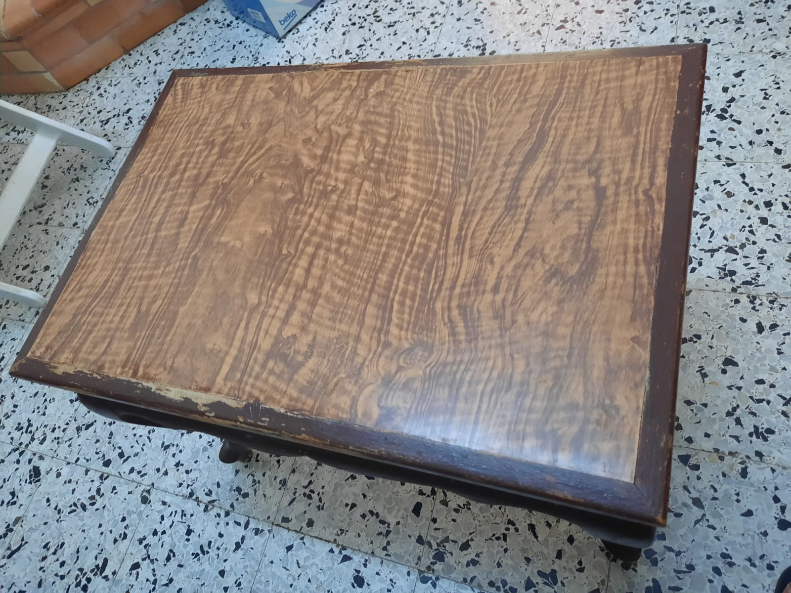 a highangle shot of a wooden table on a tiled floor. the table has a distinct wood grain pattern on its surface and a dark brown frame. the floor is covered in small, dark tiles with light grout. a white stool and a box are visible in the background.