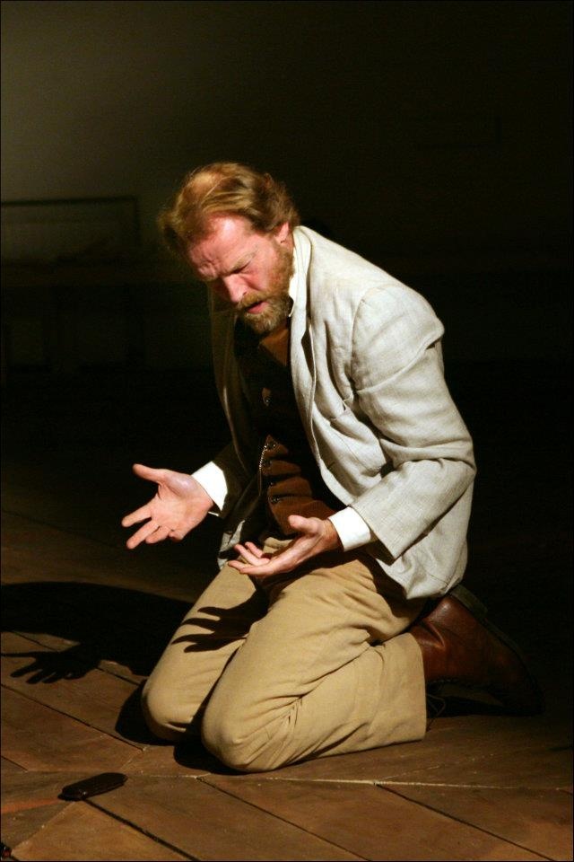 a man in a white jacket and brown pants is kneeling on a wooden floor with his head bowed and hands outstretched, as if in prayer. he is bathed in a pool of light, with his shadow cast behind him. this image evokes a sense of intense emotion, perhaps desperation or pleading. the scene appears to be set on a stage, suggesting a theatrical performance. the man's posture and the dramatic lighting contribute to a powerful and moving image.