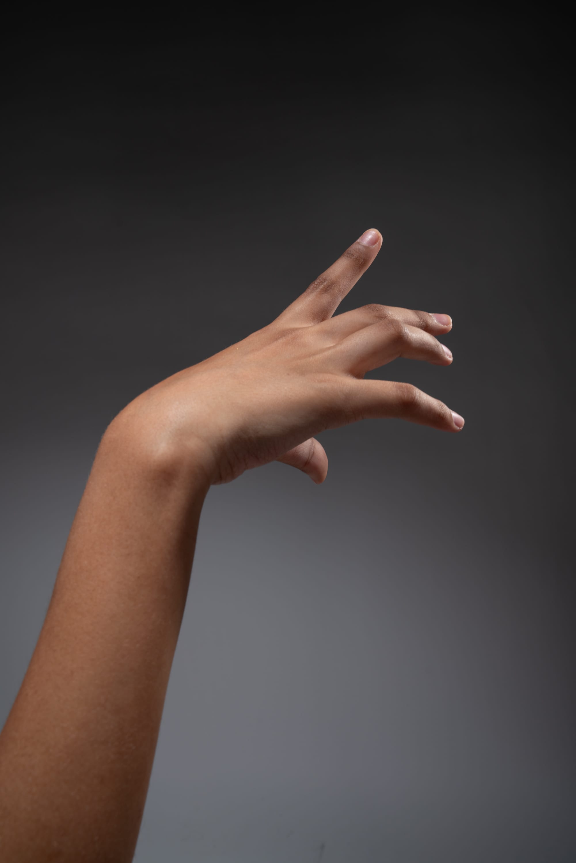 the image shows a hand with a medium brown skin tone reaching forward with the index finger extended. the background is a neutral gray and the hand is lit with studio lighting.