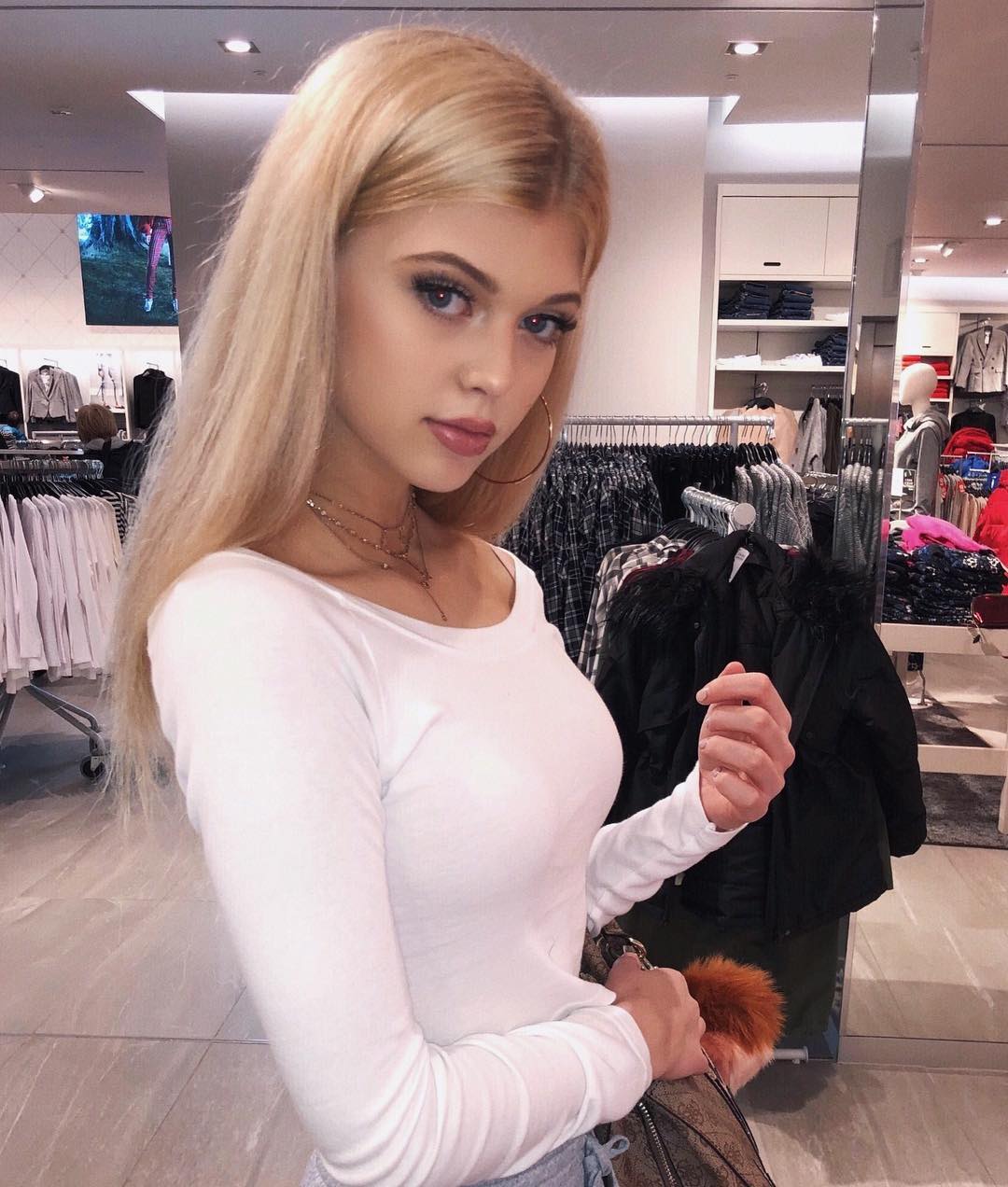 a young woman with blonde hair is standing in a clothing store, wearing a white long sleeve shirt. she is holding a handbag and is surrounded by clothing racks with various garments. the store setting includes racks of clothes, mannequins, and general retail fixtures.