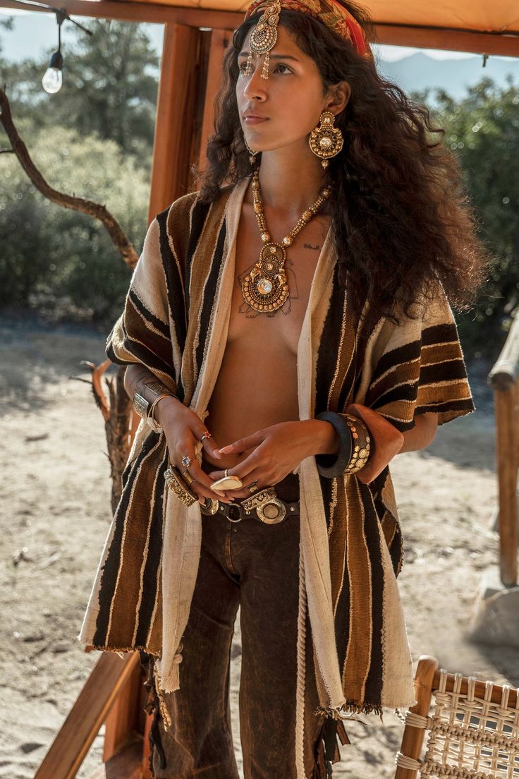 a woman with long, dark curly hair wears a beige and brown striped poncho and numerous gold jewelry pieces, including a large necklace, earrings, bracelets and a headpiece. she is standing outdoors in what looks like a desert or arid landscape, with a relaxed and serene expression on her face. the overall style is bohemian and mystical.  the setting suggests a relaxed, perhaps spiritual, atmosphere.