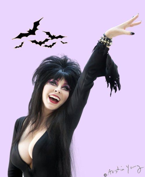a woman wearing a black dress and long black hair poses with a spooky, witchy attitude as black bats fly overhead. she is smiling with a slight wink, holding her right hand up as if to beckon someone toward her.