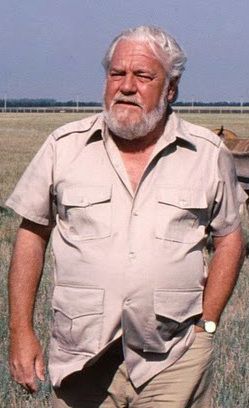 a photo of an older man standing in a field. the man has white hair and a white beard. he is wearing a short sleeve button up shirt and khaki pants. he has a watch on his left wrist and his hand is in his pocket. the background is a field.