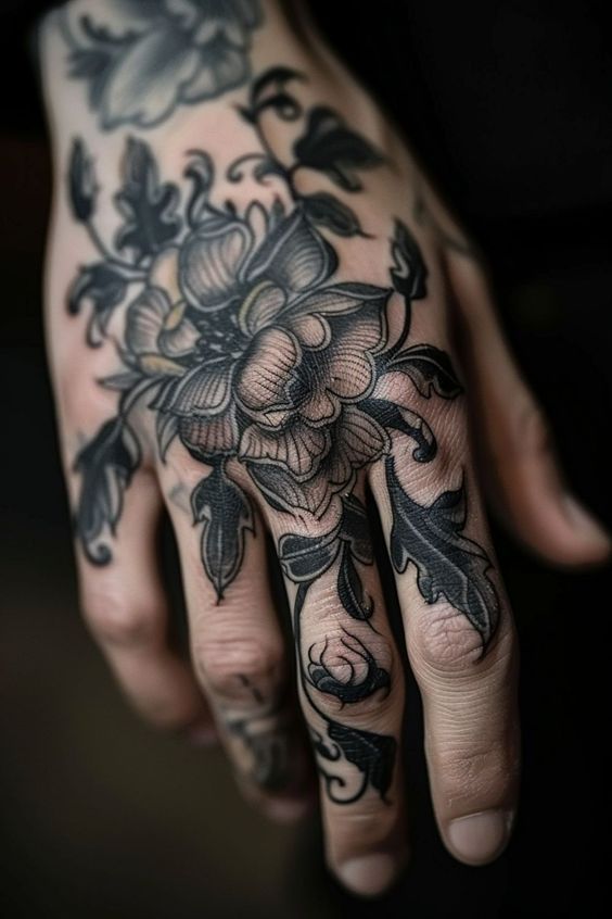a closeup image shows a hand with a detailed black and grey floral tattoo. the tattoo features a large flower and various leaves, covering the back of the hand and extending onto the fingers. the ink work is intricate and the design is botanical in nature.