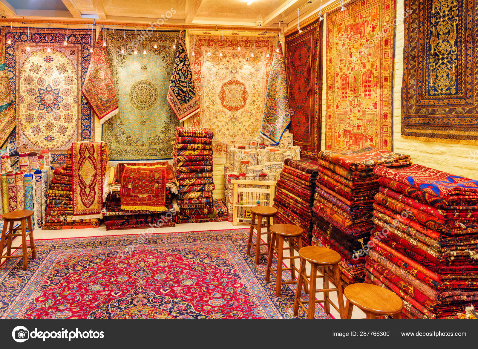 this image depicts a shop or store filled with various oriental rugs and carpets. the rugs are displayed hanging on the walls, stacked on the floor, and even used as floor coverings. the rugs feature intricate patterns, rich colors, and diverse textures. several wooden stools are positioned in the foreground. the scene is brightly lit, showcasing the vibrant textiles and creating a traditional bazaar atmosphere.