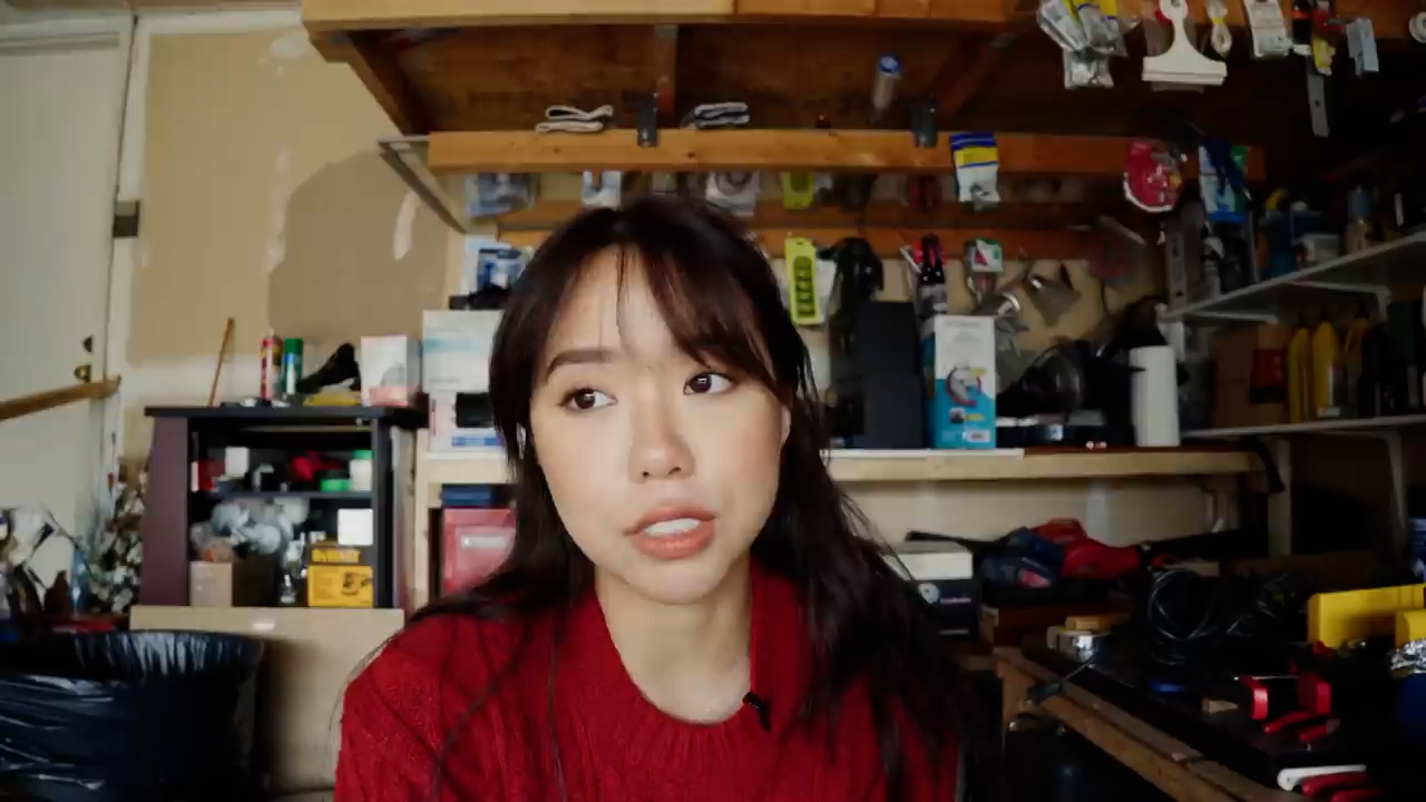 a young woman with long black hair and wearing a red sweater is standing in a garage or workshop. she is looking at the camera with a serious expression. the garage is cluttered with tools and supplies, which are organized on shelves behind her. the image appears to be a still from a video blog or tutorial about diy projects or home improvement.