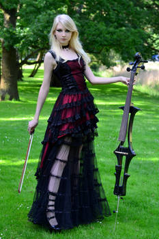a blonde woman in a gothicstyle black and red dress stands in a green park, holding an electric cello.  she is posed elegantly, and the image has a dark, romantic feel.  the dress is layered and features lace details. the cello is black and modern in design. the background is blurred, focusing attention on the woman and her instrument.