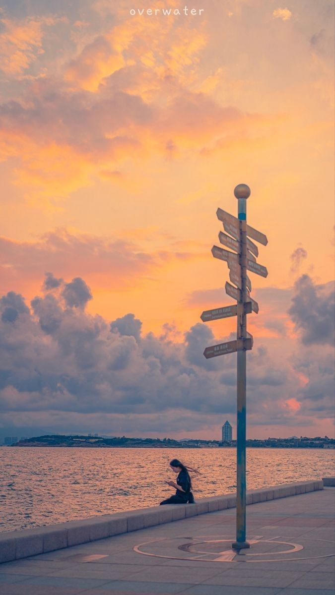 a woman sits on a seaside wall next to a signpost during sunset. the sky is filled with orange and gray clouds.