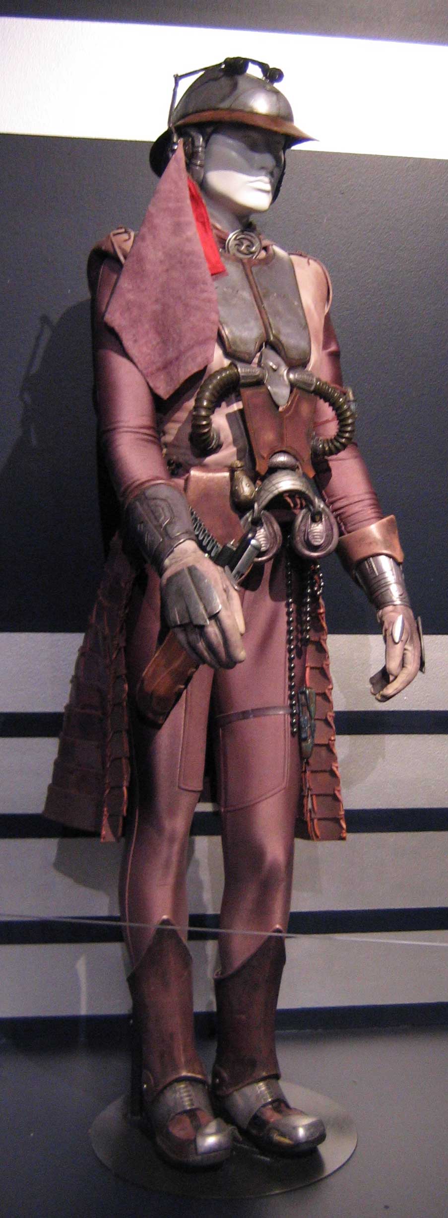 this is a detailed photo of a scifi soldier, a fantasy warrior, or perhaps a cosplay character. the figure is wearing a metallic helmet with a red sash hanging down their back, and is holding a gun in their left hand. they also appear to have a large weapon strapped to their back, possibly a blaster or a rocket launcher. the costume is very detailed, featuring buckles and other decorative elements, and the character is standing with one hand on their hip,  looking to their left. the figure is standing on a display platform, and the background appears to be a dark wall with stripes.