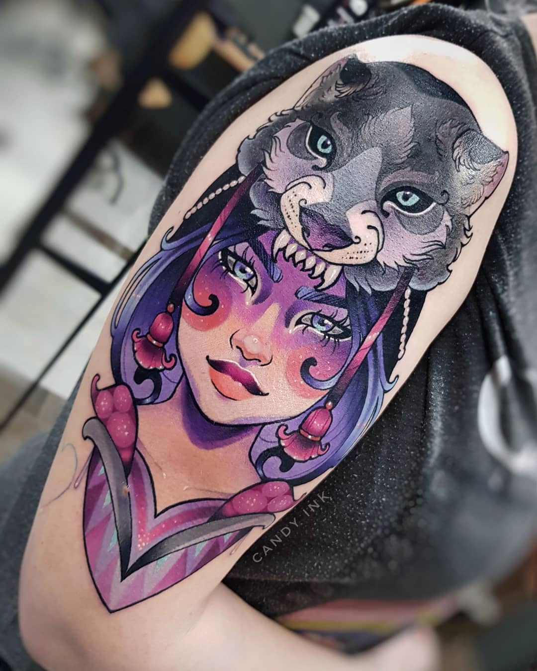 the image shows a colorful neotraditional tattoo of a woman wearing a wolf headdress. the tattoo is on the upper arm of a person. the woman in the tattoo has purple hair and blue eyes. the wolf headdress is gray with blue eyes. the tattoo is detailed and realistic, with a painterly style. the tattoo artist's signature, "candy ink", is visible below the woman's chin.