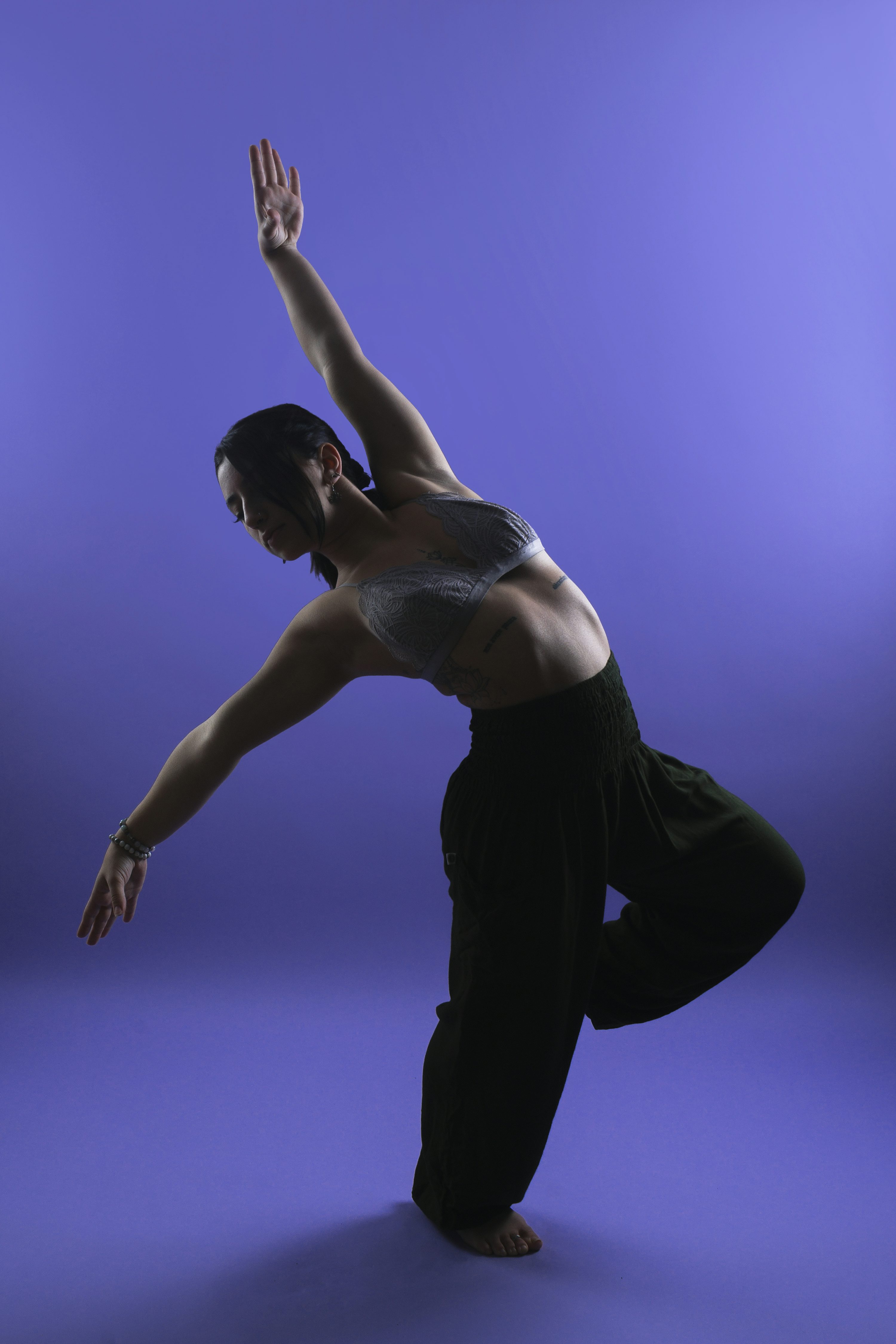 a woman is captured in a dynamic pose against a solid purple backdrop. her body is angled, suggesting motion and fluidity.  she's wearing a darkcolored bra and loosefitting pants, typical of athletic or dance attire. her hair is dark and pulled back. the lighting accentuates her form, creating a striking silhouette against the background. the overall image conveys a sense of strength, flexibility, and artistic expression.