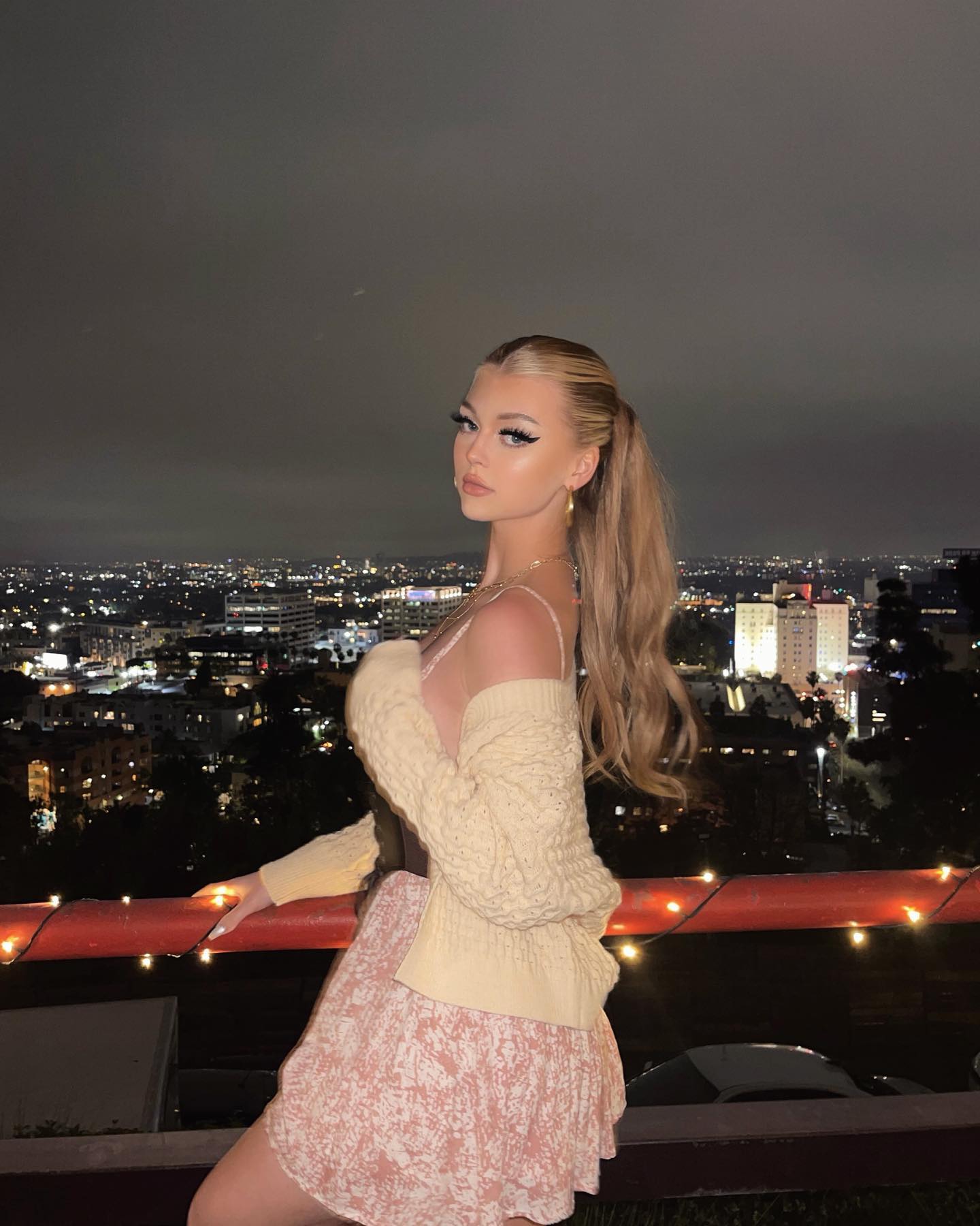 this image shows a woman named loraine standing in front of a railing with city lights in the background. she is wearing a light pink dress with a floral pattern, and a yellow cardigan. she also has a ponytail hairstyle.