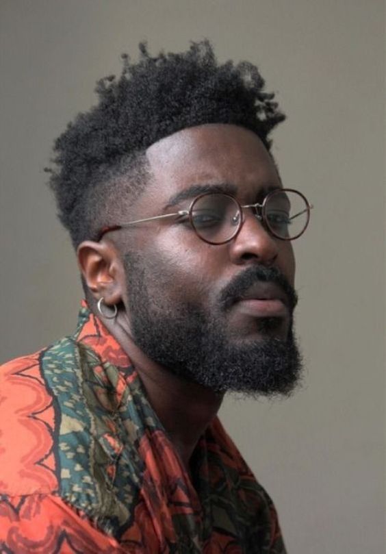 a portrait of a black man wearing round glasses, a beard, and an earring. he has a fade hairstyle. he is wearing a colorful, patterned shirt.
