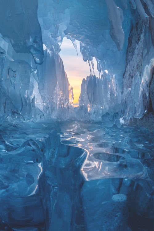the image shows a frozen ice cave with sunlight shining through the opening. the ice is a translucent blue color and the cave is filled with icicles and frozen formations. the ground is covered in ice and there is a hint of a landscape visible through the cave opening.