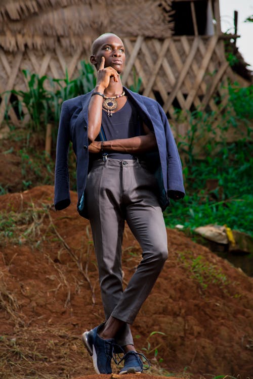 the image shows a bald, darkskinned male model standing outdoors in a rural african setting. he's dressed in a stylish outfit:  a dark blue blazer worn casually over a dark shirt and gray dress pants. he's wearing sneakers and a distinctive necklace. his pose is contemplative, with his hand to his chin, looking thoughtfully upward. the background features a traditional thatched hut and lush vegetation, creating a striking contrast with the model's modern attire. the overall impression is one of sophisticated fashion photography against a rustic backdrop.