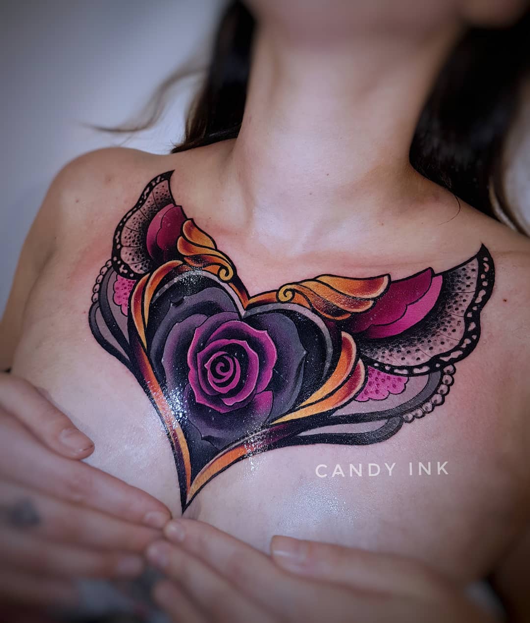 a closeup photograph showcasing a freshly done chest tattoo on a woman. the tattoo features a heartshaped design with a black and purple rose in the center. the heart is adorned with golden wings and intricate lace patterns in shades of pink and grey. the tattoo artist's watermark "candy ink" is subtly placed at the bottom center. the woman's hands gently touch the top of her chest, drawing attention to the new ink.  the soft, artificial lighting enhances the vibrant colors of the tattoo.