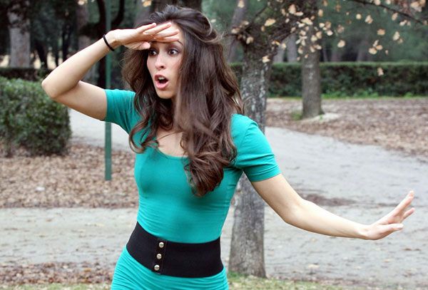 a young woman in a teal dress is standing in a park, looking surprised and pointing with her hand. she is wearing a black belt and has long brown hair. the background is a park with trees and a path. the lighting is natural and overcast.