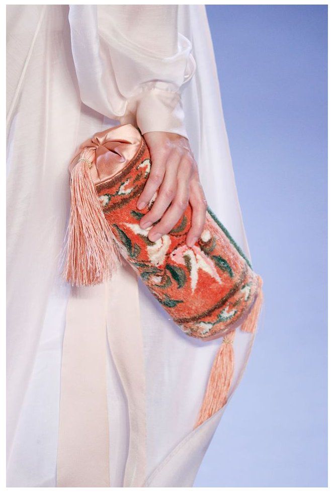 closeup view of a model holding a unique cylindrical clutch bag. the clutch is made of a richly patterned fabric resembling a carpet or rug, with predominantly orange and green floral motifs. it's adorned with delicate pink silk tassels.  the model is wearing a flowing white silk dress, suggesting a high fashion context such as a runway show. the image emphasizes the texture and detail of the clutch against the smooth simplicity of the dress.