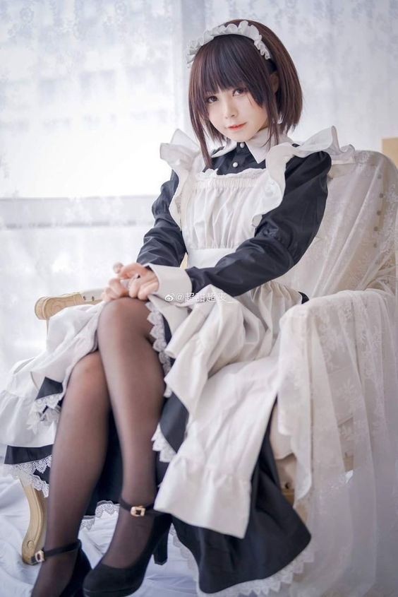 a young asian woman in a maid costume is sitting in a chair in a photo shoot. she is wearing a black and white dress, black stockings, and black shoes. the photo is taken from eye level and uses studio lighting.