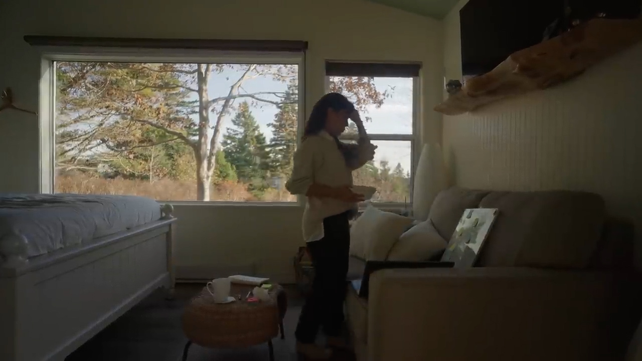 a woman stands in profile in a naturally lit bedroom, facing a window with a view of trees and nature. she holds a bowl in one hand, suggesting she is eating, and has her other hand on her head. the room features a bed on the left, a couch on the right with a laptop resting on it, and a small table with a mug and other items. the overall atmosphere is calm and relaxed.