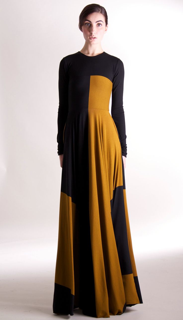 a woman is standing in a studio wearing a long, black and gold dress. the dress has long sleeves and a fitted bodice. the woman is looking at the camera with a serious expression.
