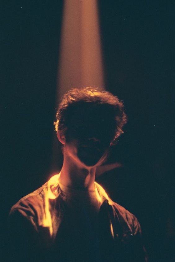 a man stands on a dark stage illuminated only by a single, strong spotlight from above. his face is obscured by shadow, creating a sense of mystery and drama.  the lighting emphasizes the contours of his face and upper body, highlighting his presence in the otherwise dark environment.