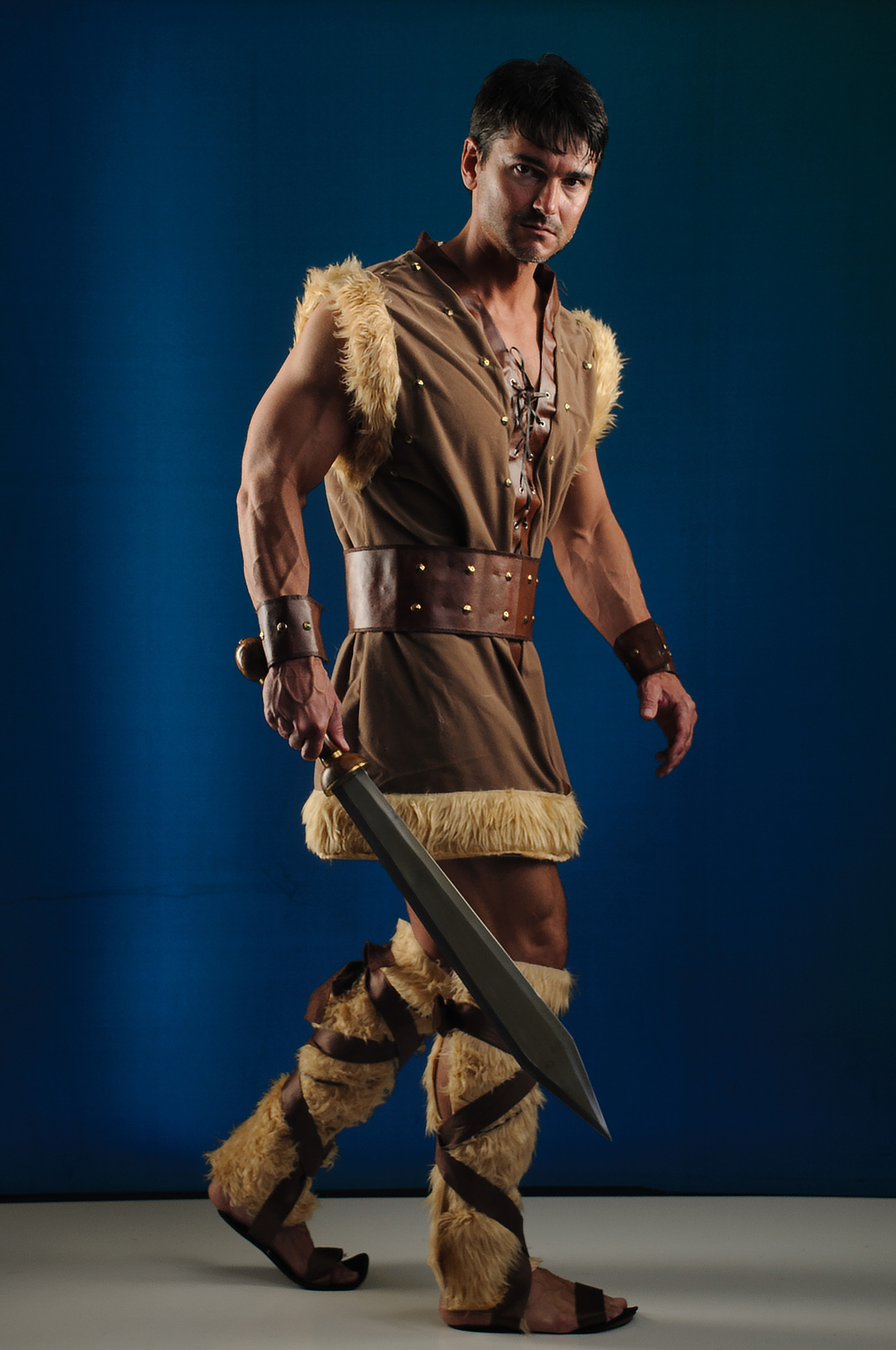 a muscular man dressed in a warrior costume, holding a sword, poses against a blue background. he is wearing a furtrimmed tunic, leather belt, wristbands, and leg warmers, along with sandals. he has dark hair and a serious expression as he looks at the camera.