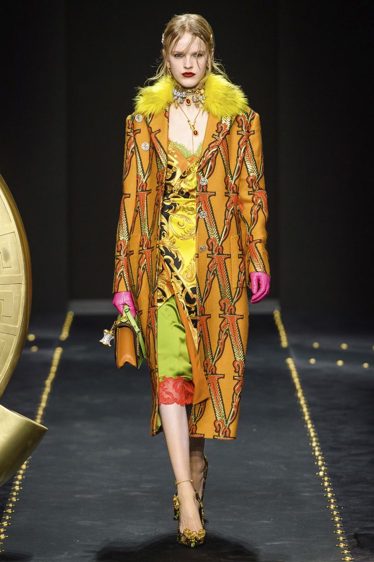a model walks down the versace runway wearing a striking ensemble.  the outfit features a long, orange coat with a large "v" pattern, layered over a vibrant yellow and gold slip dress with a green and red underlay.  she also sports a yellow fur collar, pink gloves, gold jewelry including a choker and necklaces, and ornate gold heels. the overall look is a bold mix of colors and textures, indicative of high fashion. the background suggests a dark runway with gold accents.