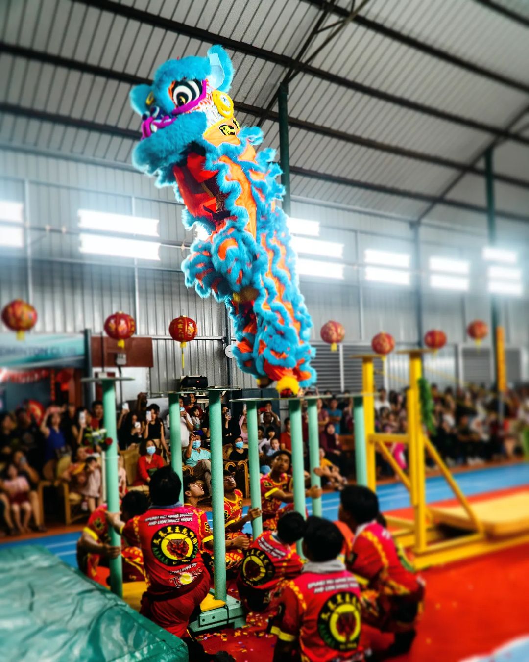 a vibrant lion dance troupe performs atop poles, their colorful costume a blur of blue, orange, and yellow against the bright lights of an indoor gymnasium. an engaged audience watches and captures the spectacle on their phones. the atmosphere is festive, likely in celebration of chinese new year.