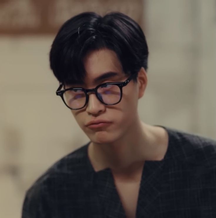 a young man with black hair and glasses is pictured in a medium closeup shot. he is wearing a darkcolored shirt with a loose neckline. he appears to have a neutral or slightly annoyed expression. the lighting is soft, creating a natural and approachable look. the background is blurred, suggesting an indoor setting.