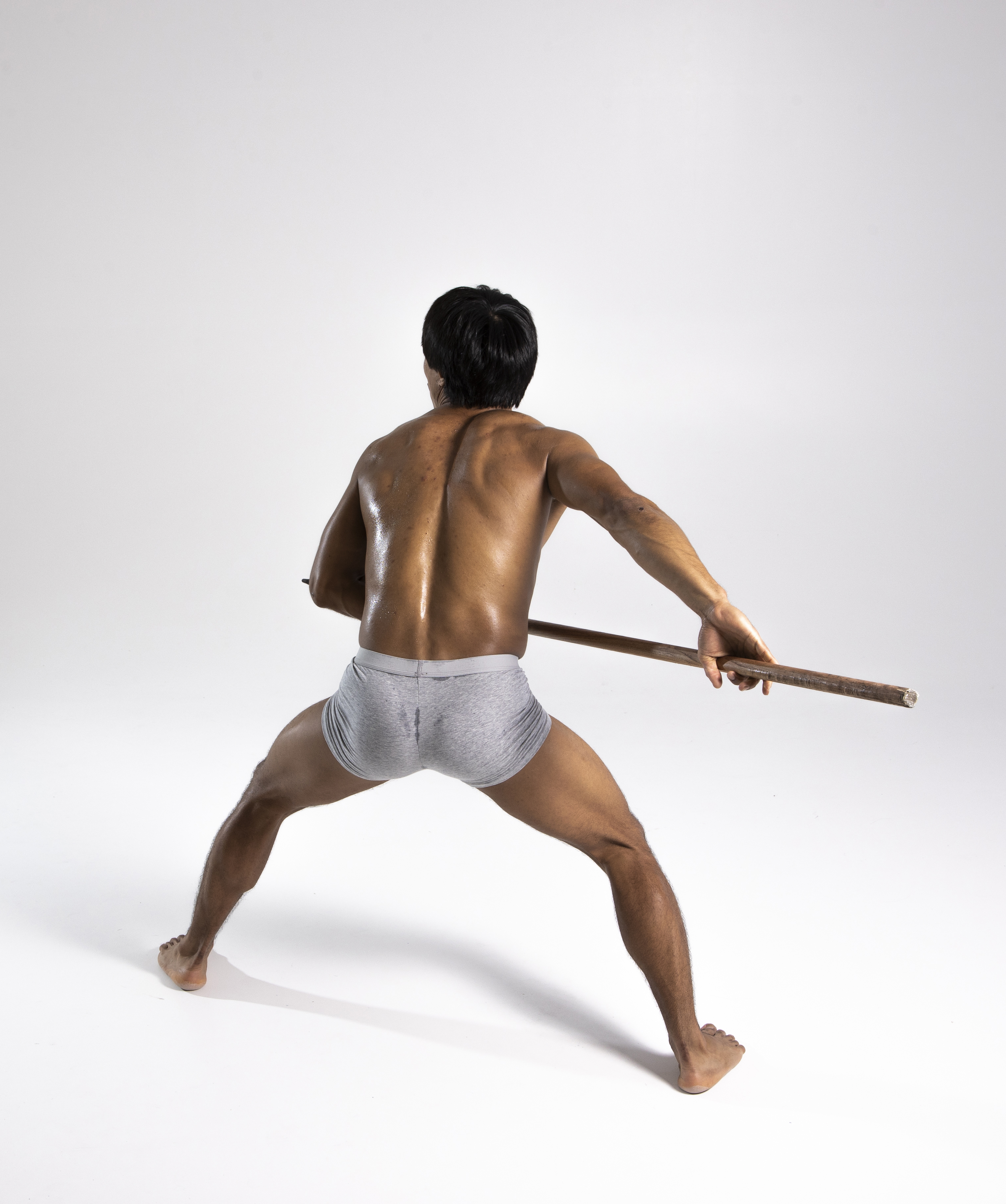 a shirtless, muscular man with dark hair is standing with a wide stance, holding a wooden stick behind his back. he is wearing gray shorts. the photo is a studio shot against a white background. he appears sweaty. his back is to the viewer.