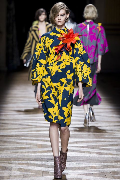the image shows a fashion runway show. the focus is on a central model wearing a striking kneelength dress with a bold yellow and blue floral print. the dress features a high neckline and wide sleeves. the model also wears ankle boots and has a large, vibrant red and orange flower brooch pinned to her dress.  in the background, two other models are visible, also showcasing distinct fashion styles; one in a striped jacket, the other in fuchsia and grey.  the setting appears to be a wooden runway, suggesting a professional fashion show environment. the overall mood is chic and sophisticated.