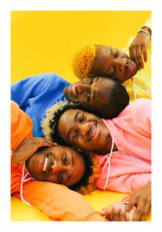 four young black people are  laying closely, laughing and smiling at the camera against a bright yellow backdrop.  they are wearing brightly colored hoodies in orange, pink, blue, and yellow. the image evokes feelings of joy, friendship, and diversity.