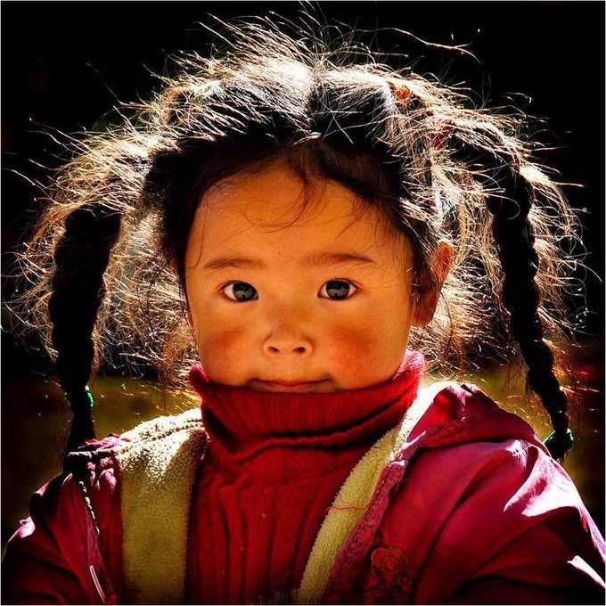 a closeup portrait of a young girl with her hair styled in pigtail braids. she is wearing a red turtleneck sweater and a pink jacket with a lightcolored lining. her cheeks are rosy, and her face is lit by sunshine, creating a play of light and shadow.