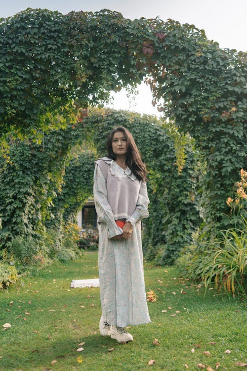 a young asian woman stands in a garden archway. she is wearing a long floral dress, a light purple sweater vest, and white ankle boots. she holds a small orange clutch in her hands. the garden is lush with green vines and plants. the overall mood is serene and peaceful.
