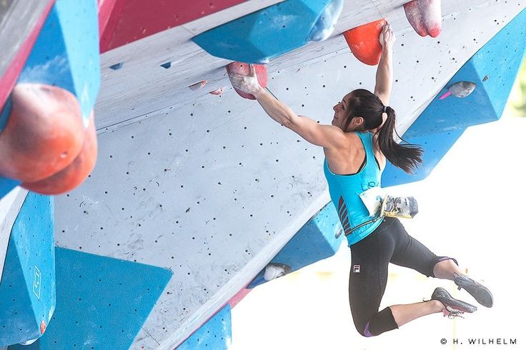 a female climber is shown suspended in midair on a challenging bouldering wall. she is seen reaching for the next hold with her right hand, her body stretched out horizontally. the vibrant colors of the climbing wall and her attire add to the energy of the scene.