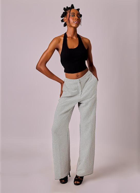 a full shot of a slender african american woman standing and looking confidently in a studio setting. she has her hands in her pockets and is wearing a black halter top, checkered highwaisted wideleg pants, and black highheeled sandals. her hair is styled in knots. the background is a plain light color.