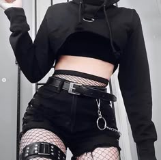 a young woman is dressed in a black crop top hoodie with a black belt and fishnet tights. she is posing for a picture. the overall style is edgy and trendy. the outfit is mostly black which gives off a cool and casual vibe.