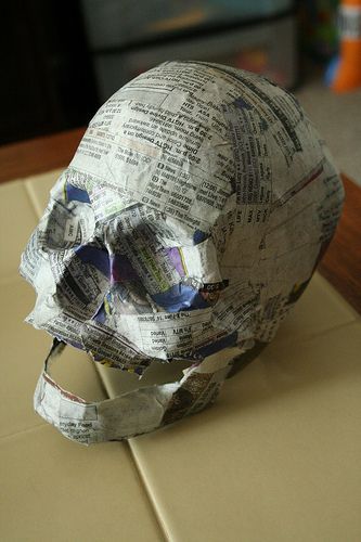 a closeup of a papiermache skull made out of newspaper. the skull is unfinished and still has the raw newspaper showing. the skull is lying on a table and there is a light beige background behind it. the skull is a craft project that could be used for halloween decorations.