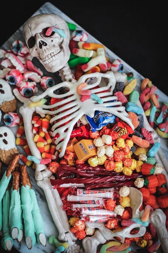 the image shows a plastic skeleton model with various halloween candies arranged within and around its ribcage and limbs.  the candies include gummy worms, candy corn, popcorn, licorice, colorful gummy candies,  eyeballs, pretzels shaped like fingers, ghostshaped cookies, and various commercially packaged candies like milk duds and reese's pieces. the overall effect is a creative and slightly macabre food arrangement perfect for a halloween party or display. the skeleton lies on a marble surface.