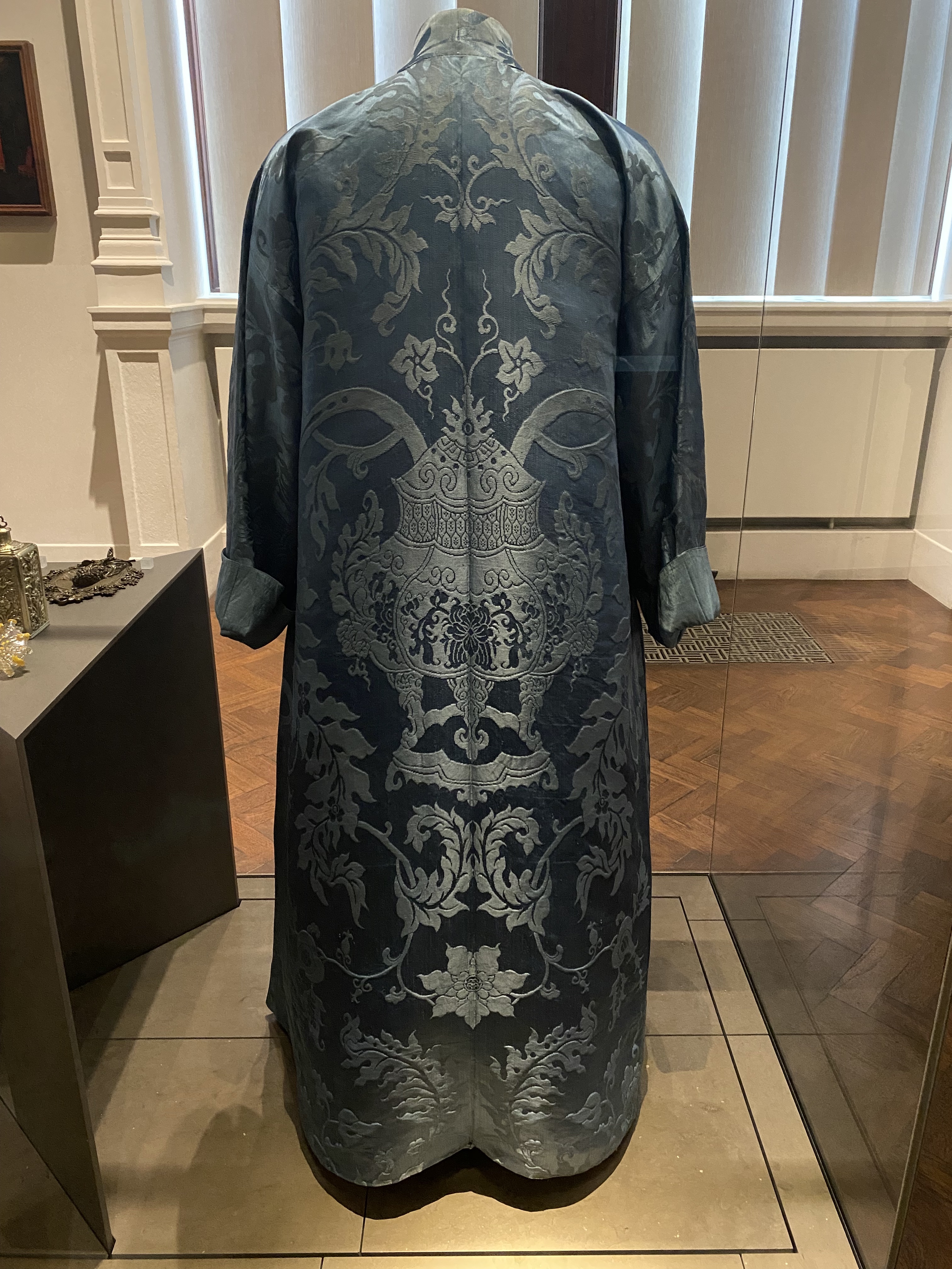 a long, blue patterned robe is displayed in a museum behind a glass display case. the robe has a complex pattern on it that appears to be floral or botanical. the robe has long sleeves and is of a chinese style.