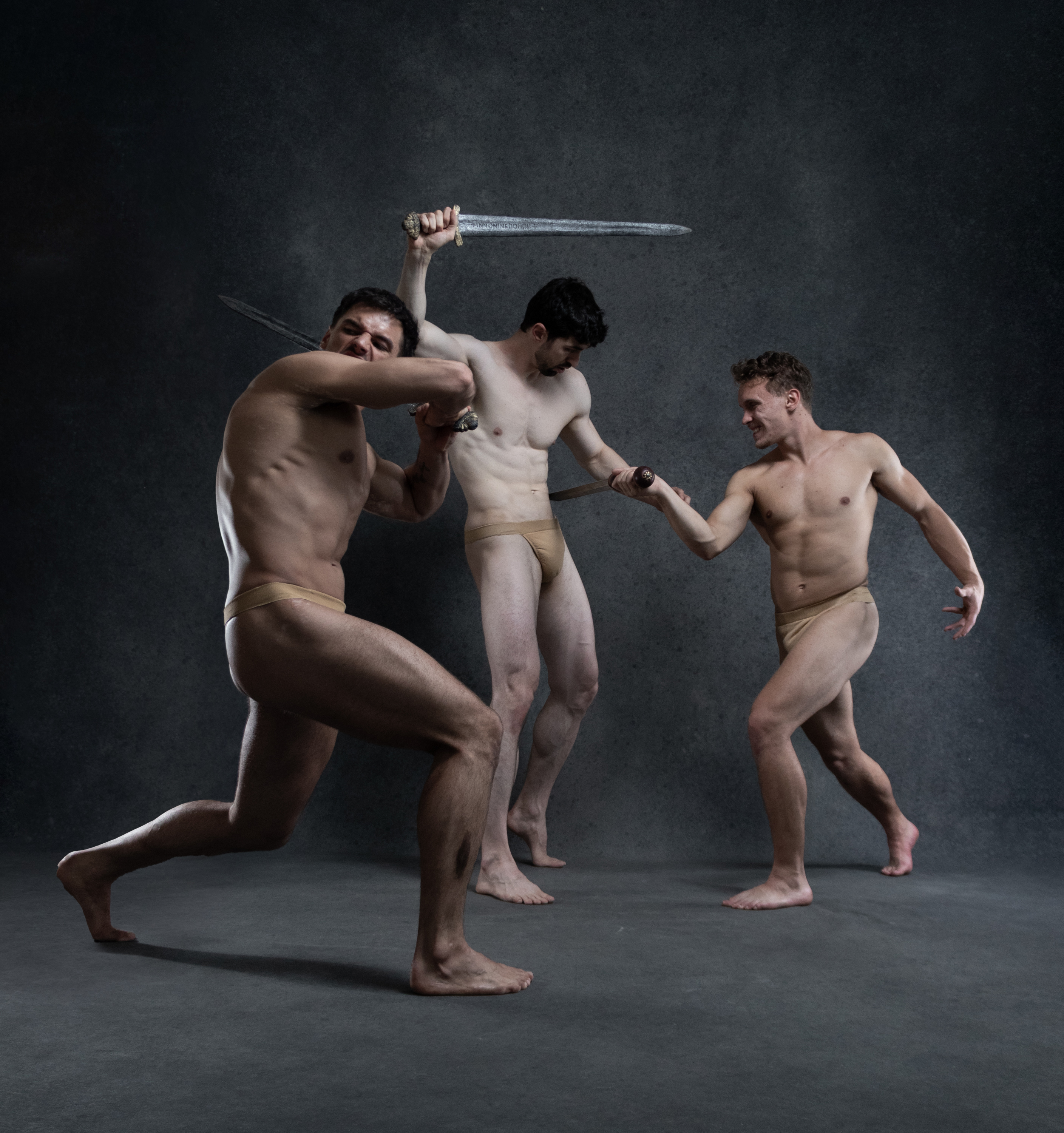 three muscular men in beige loincloths are engaged in a dynamic sword fight against a dark gray backdrop.  they are illuminated with dramatic studio lighting that highlights their physiques. the man on the left, positioned in a defensive stance, attempts to block the attack of the man on the right, who lunges towards him. in the background, another man stands with his sword raised.  the scene evokes a sense of ancient warriors engaged in fierce combat.